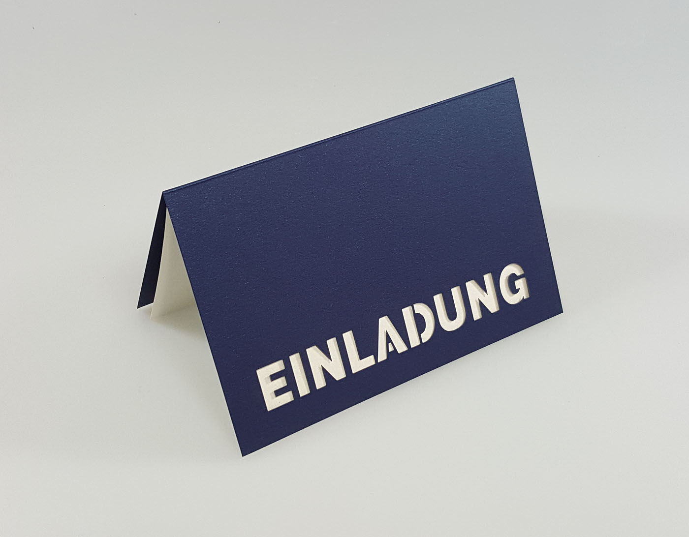 Dunkelblaue Einladungskarte mit ausgestanztem Schriftzug "EINLADUNG" und weißem Einlegeblatt - Aufgestellte Ansicht