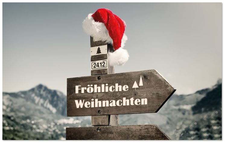 Lustiger Weihnachtsgruß mit "Fröhliche Weihnachten" auf Wegweiser in den Bergen und roter Mütze